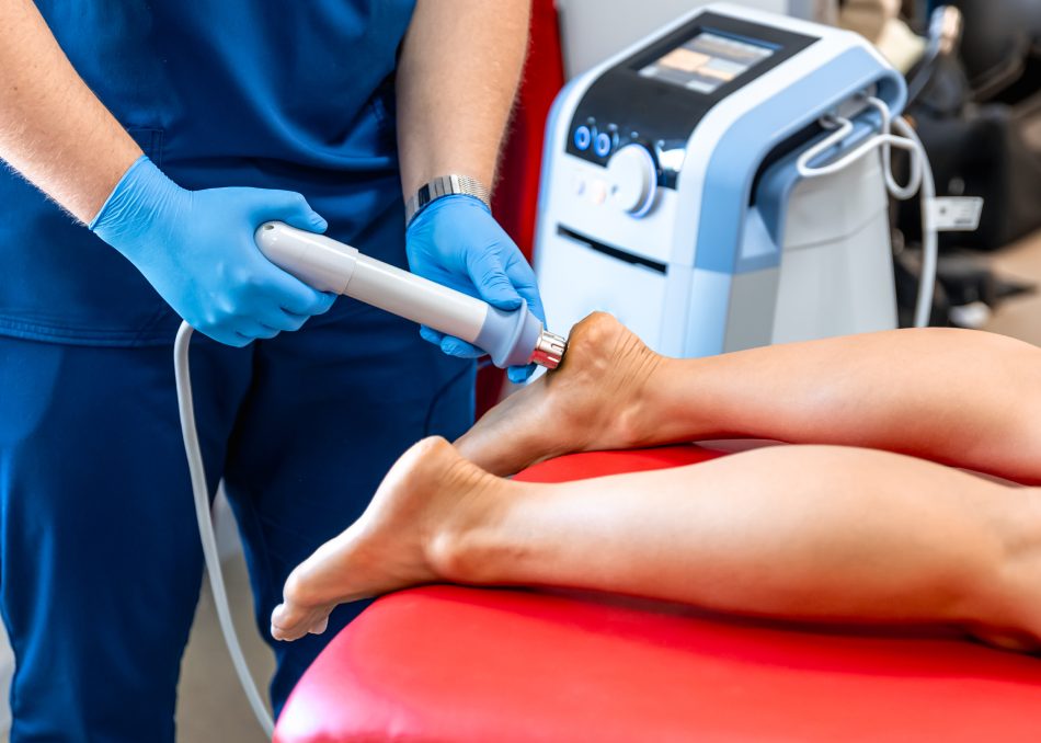 A gloved provider uses a shockwave therapy device on a patient's foot and ankle during a podiatric treatment session