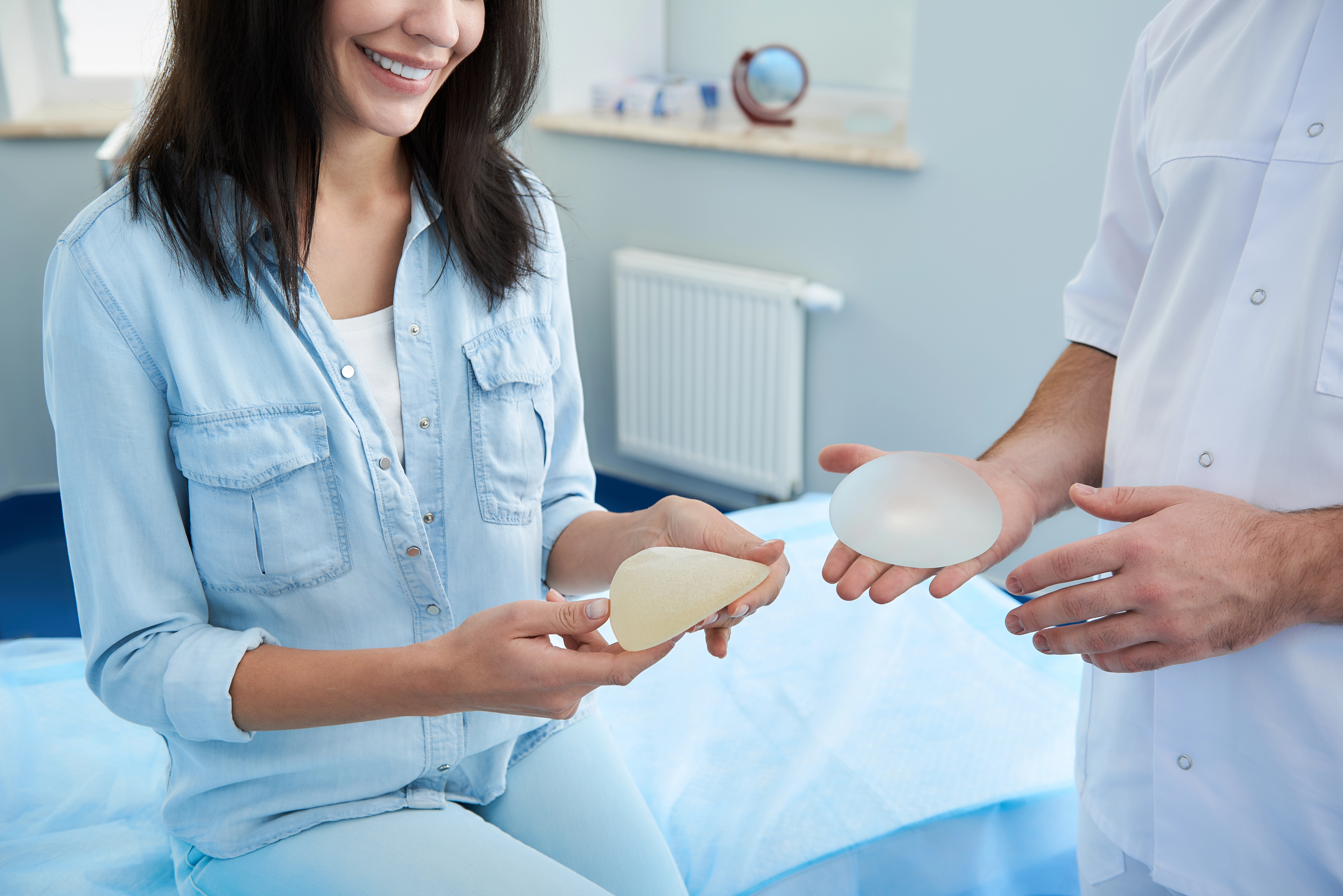 A smiling woman consults with a doctor while comparing two different breast implant sizes in an exam room