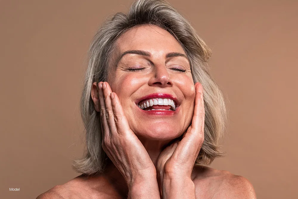 A mature woman with closed eyes smiles broadly while touching her face with both hands against a warm neutral background