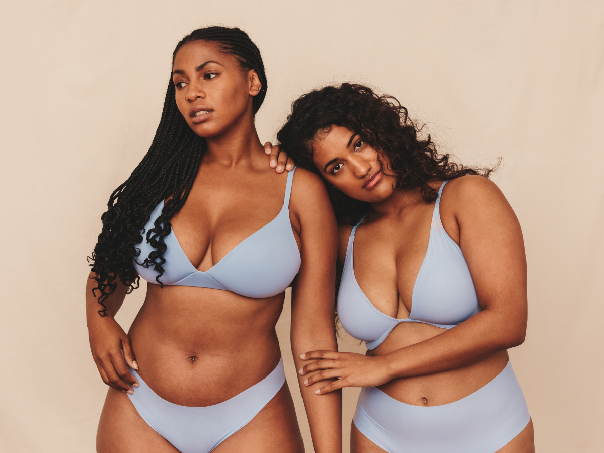 Two women in matching light blue underwear pose together against a neutral studio backdrop, showcasing natural body confidenc