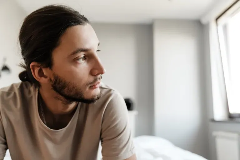 A pensive young man with long hair sits on a bed near a window, gazing off in thought, illustrating anxiety in a blog post ab