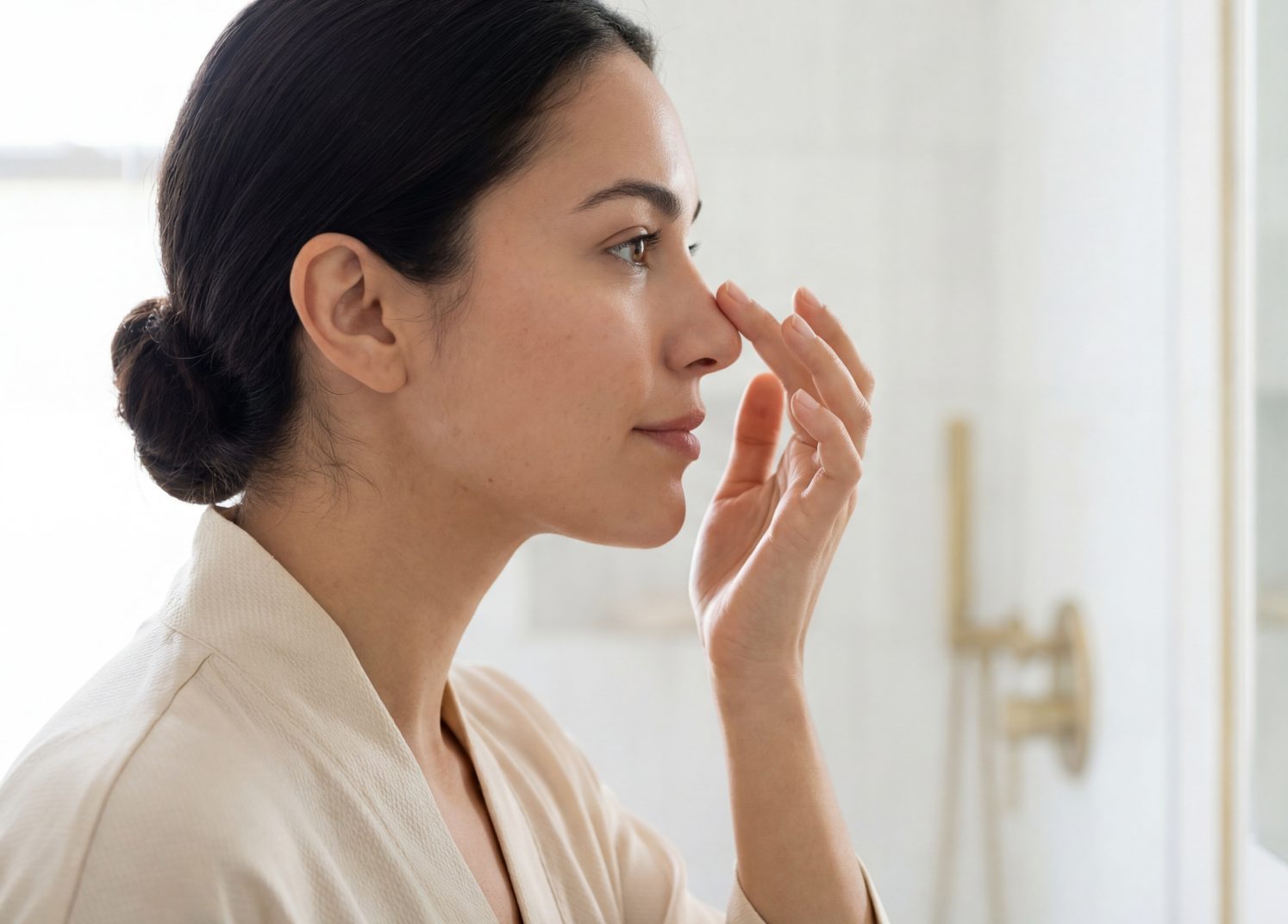 Woman in a beige robe touching the bridge of her nose while examining her profile in a bathroom mirror