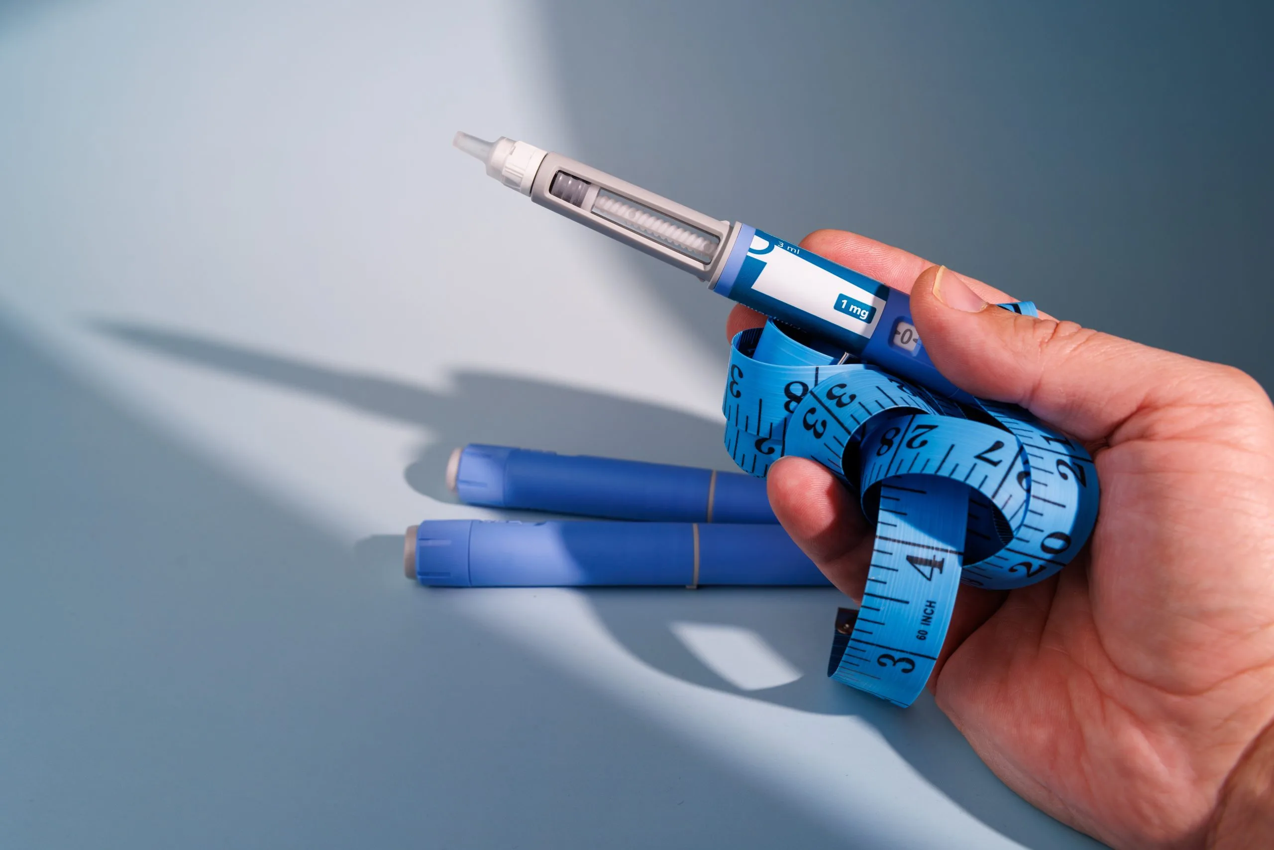 Hand holding injectable pen with blue measurement tape wrapped around it and two additional pens below