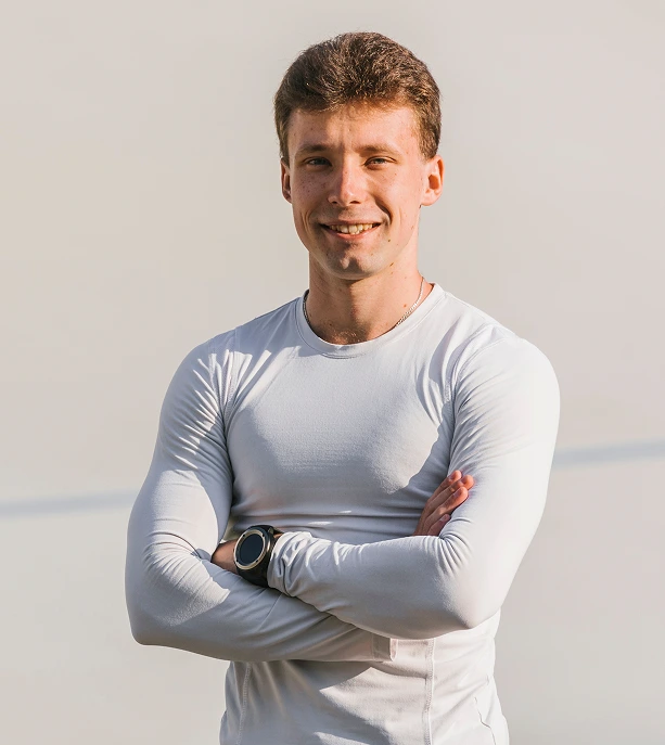 Man in light gray long-sleeve shirt standing with arms crossed, smiling at camera against neutral background