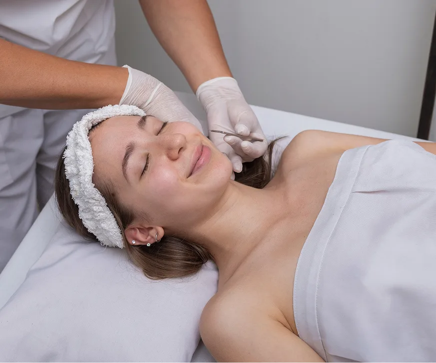 Aesthetician in white gloves performing facial treatment on relaxed patient with towel headwrap