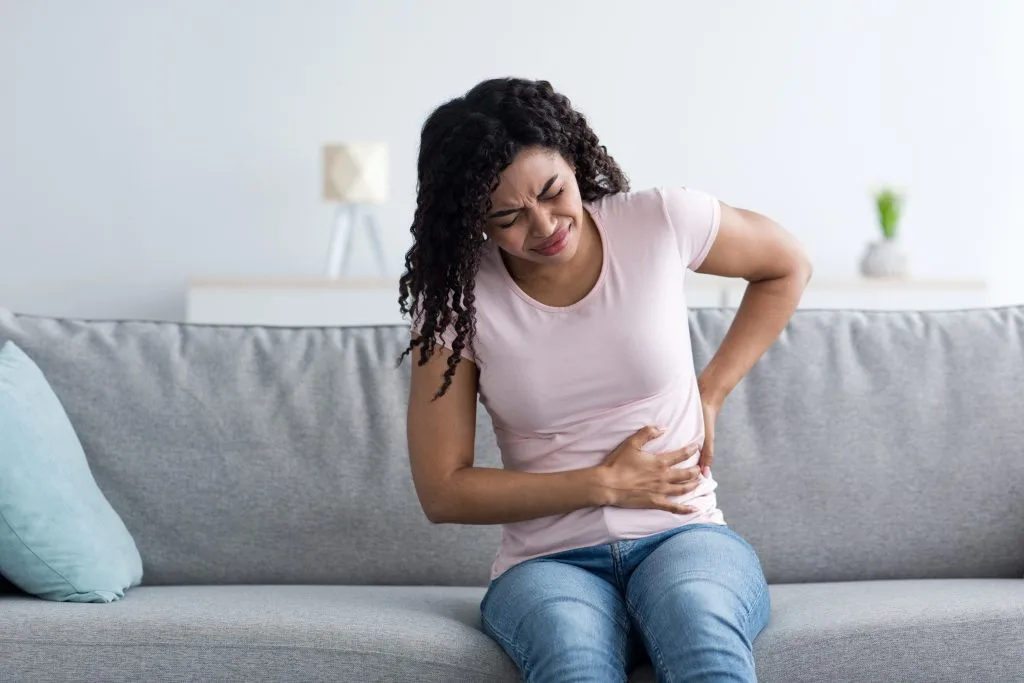 Woman sitting on couch holding her abdomen while wincing in pain