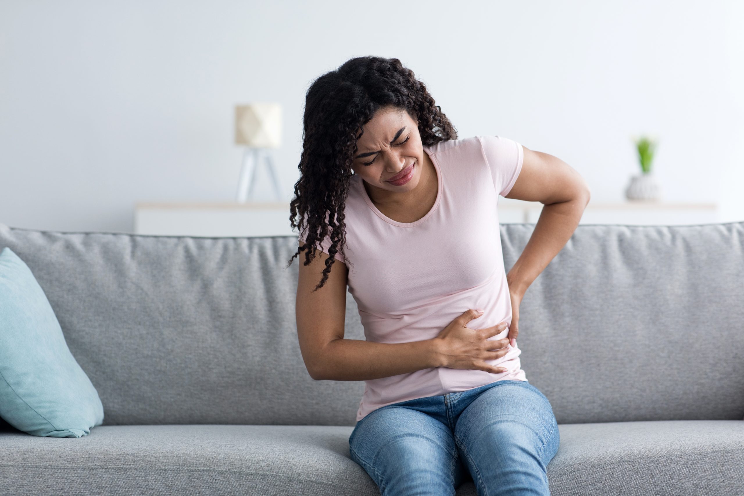 Woman sitting on couch holding her abdomen while wincing in pain