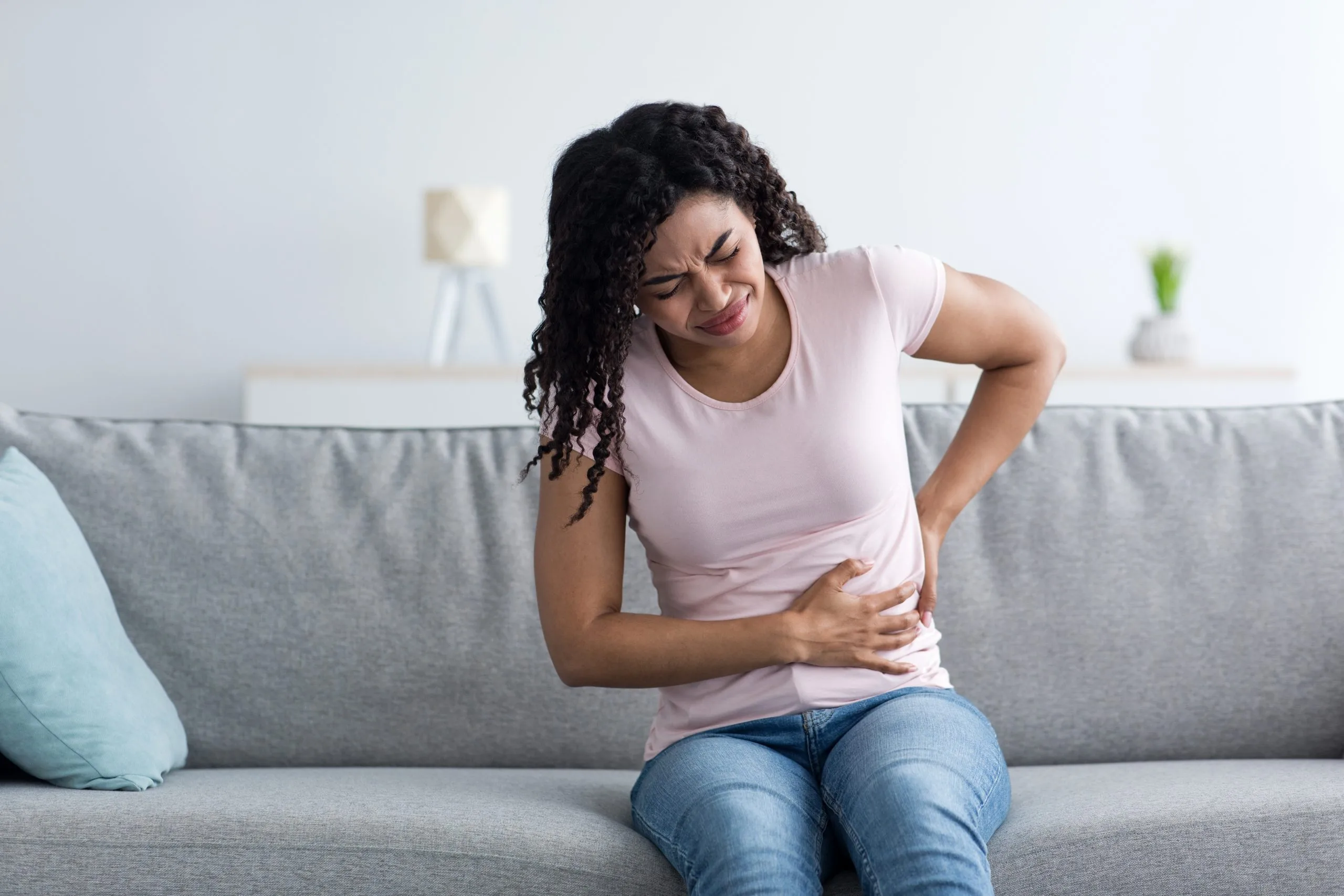 Woman sitting on couch holding her abdomen while wincing in pain