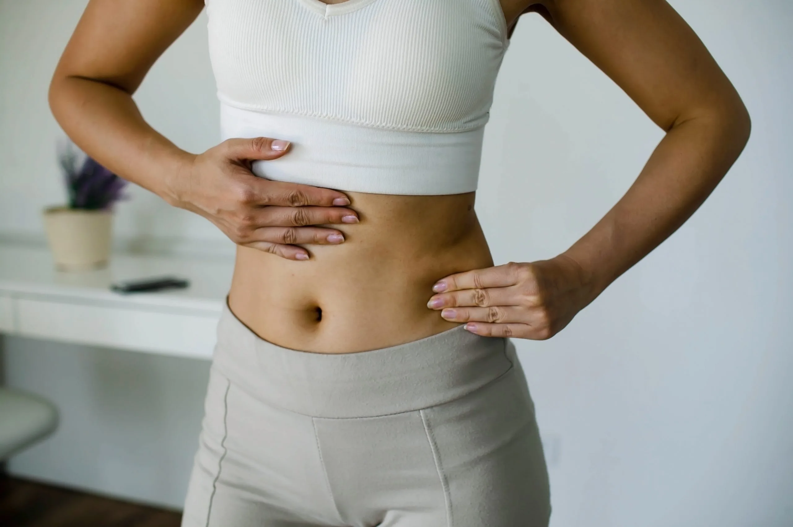 Woman in white crop top and low-rise pants touching her abdomen in a studio setting