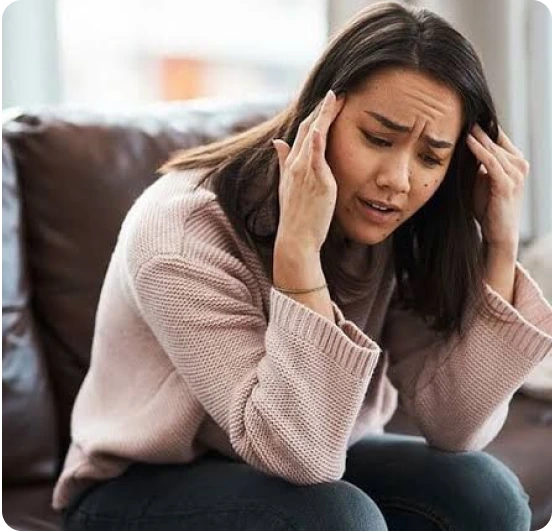 Woman sitting on couch holding her head with both hands, appearing stressed or experiencing concentration difficulties