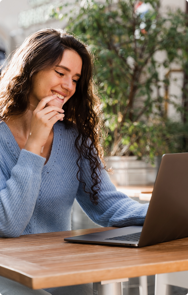 Woman smiling while using a laptop at a wooden desk with greenery in the background