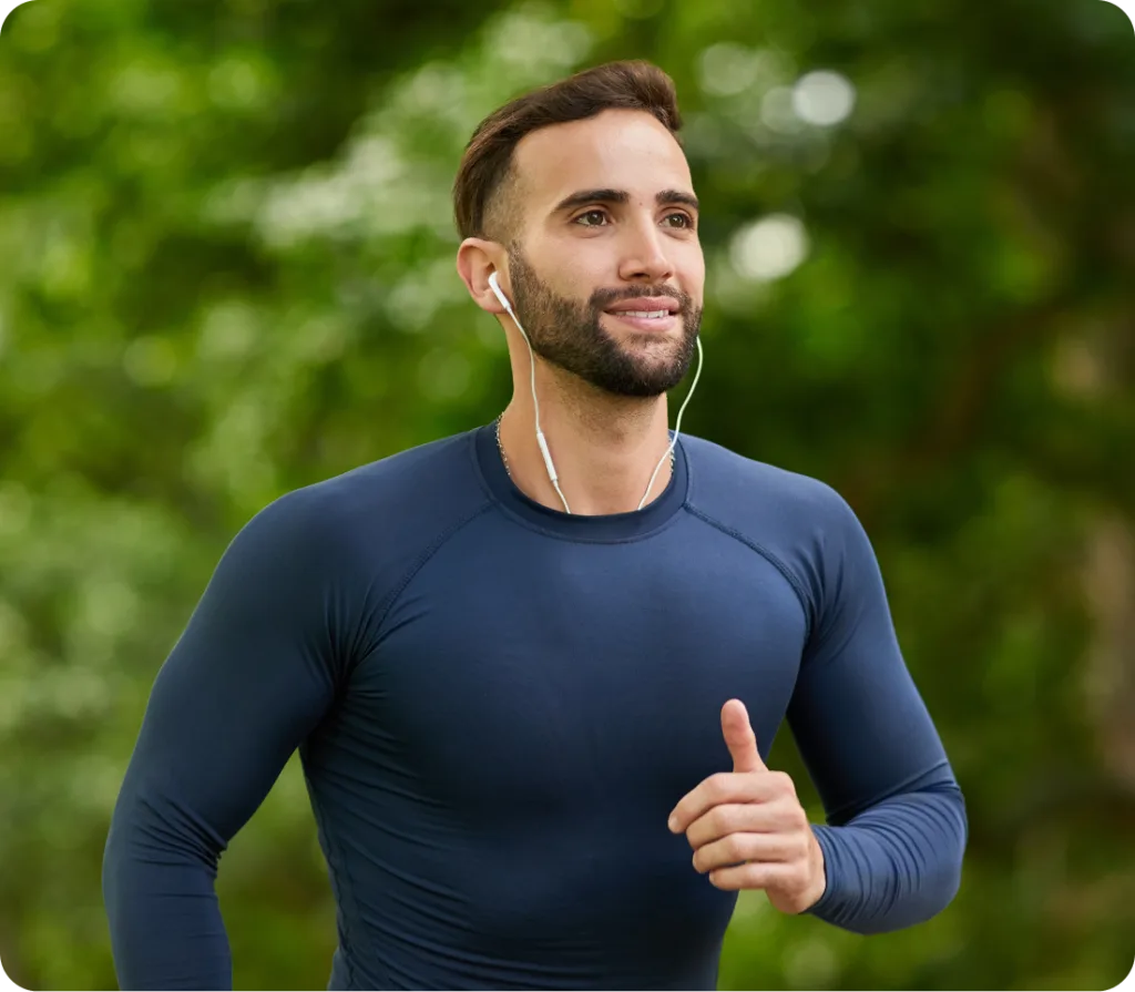 Man jogging outdoors wearing earbuds and giving thumbs up with green foliage background