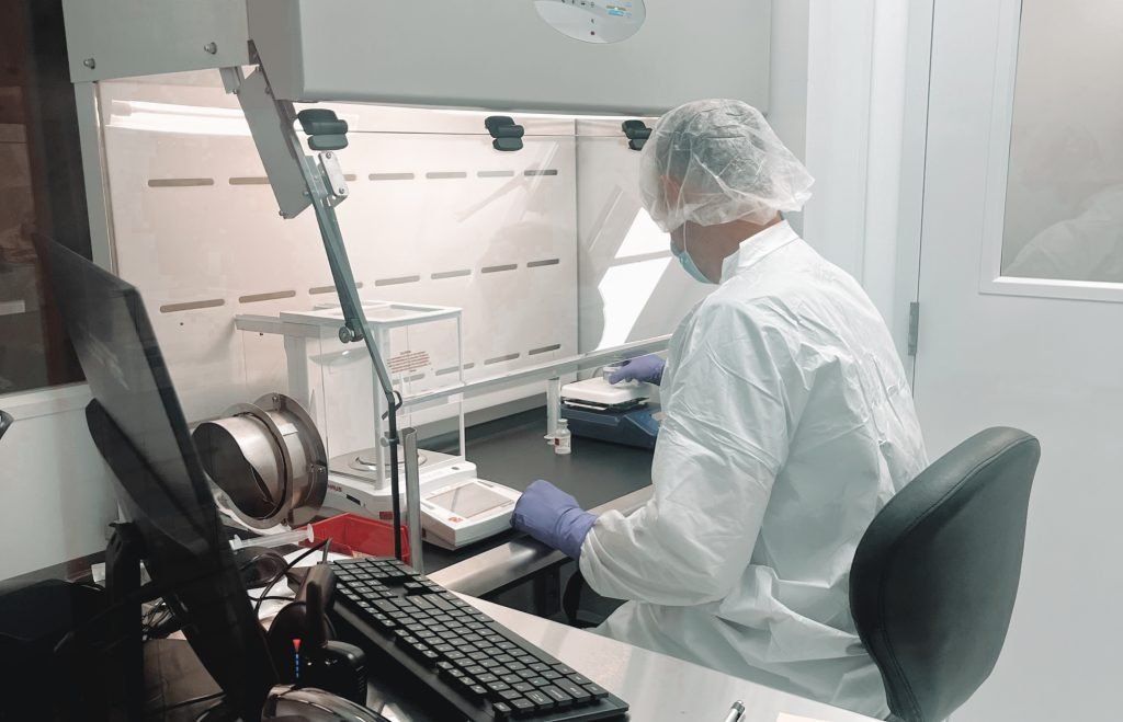 Pharmacy technician in protective gown and hair cover preparing a compounded medication at a sterile hood