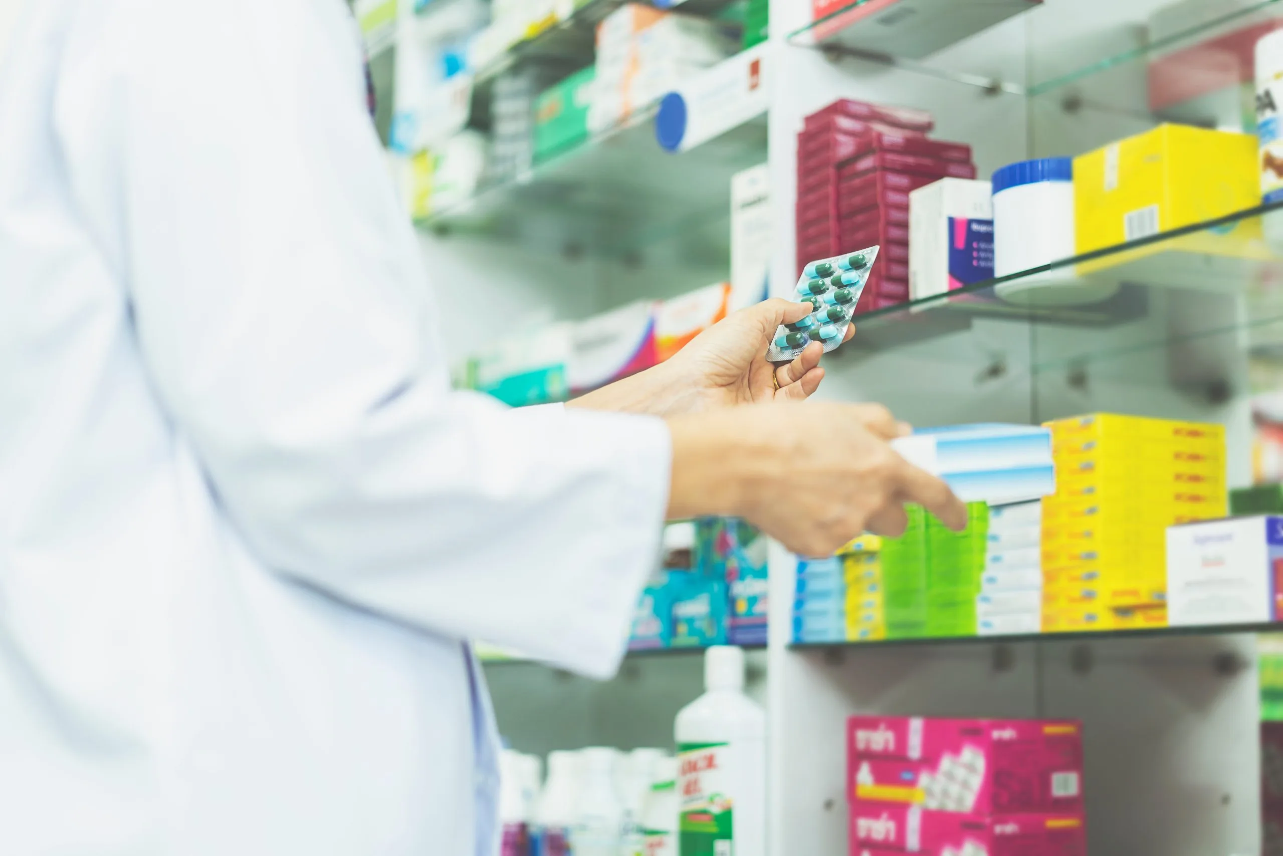Pharmacist holding a blister pack of capsules in front of pharmacy shelves stocked with colorful medicine boxes