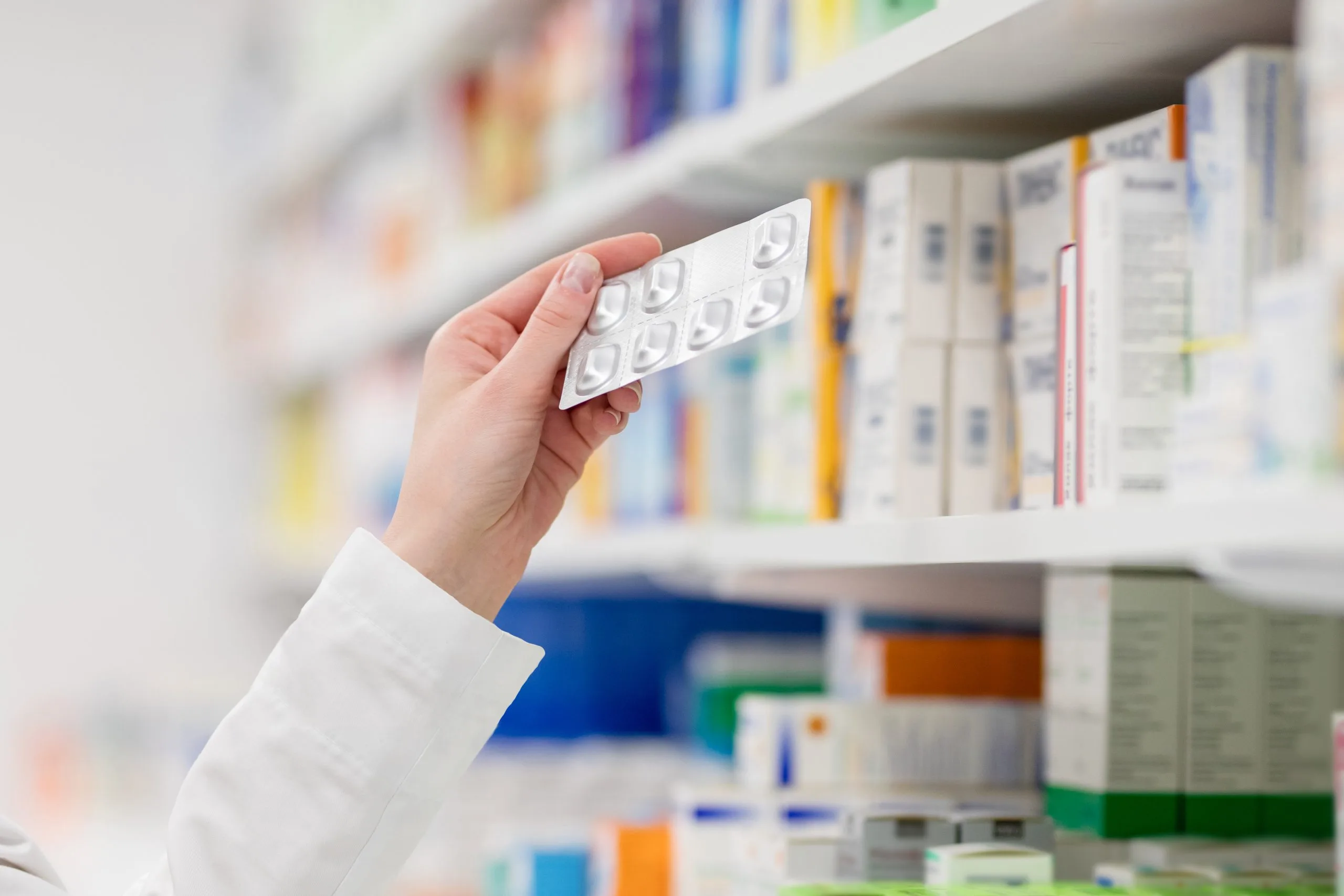 Pharmacist's hand holding a blister pack of pills in front of stocked pharmacy shelves