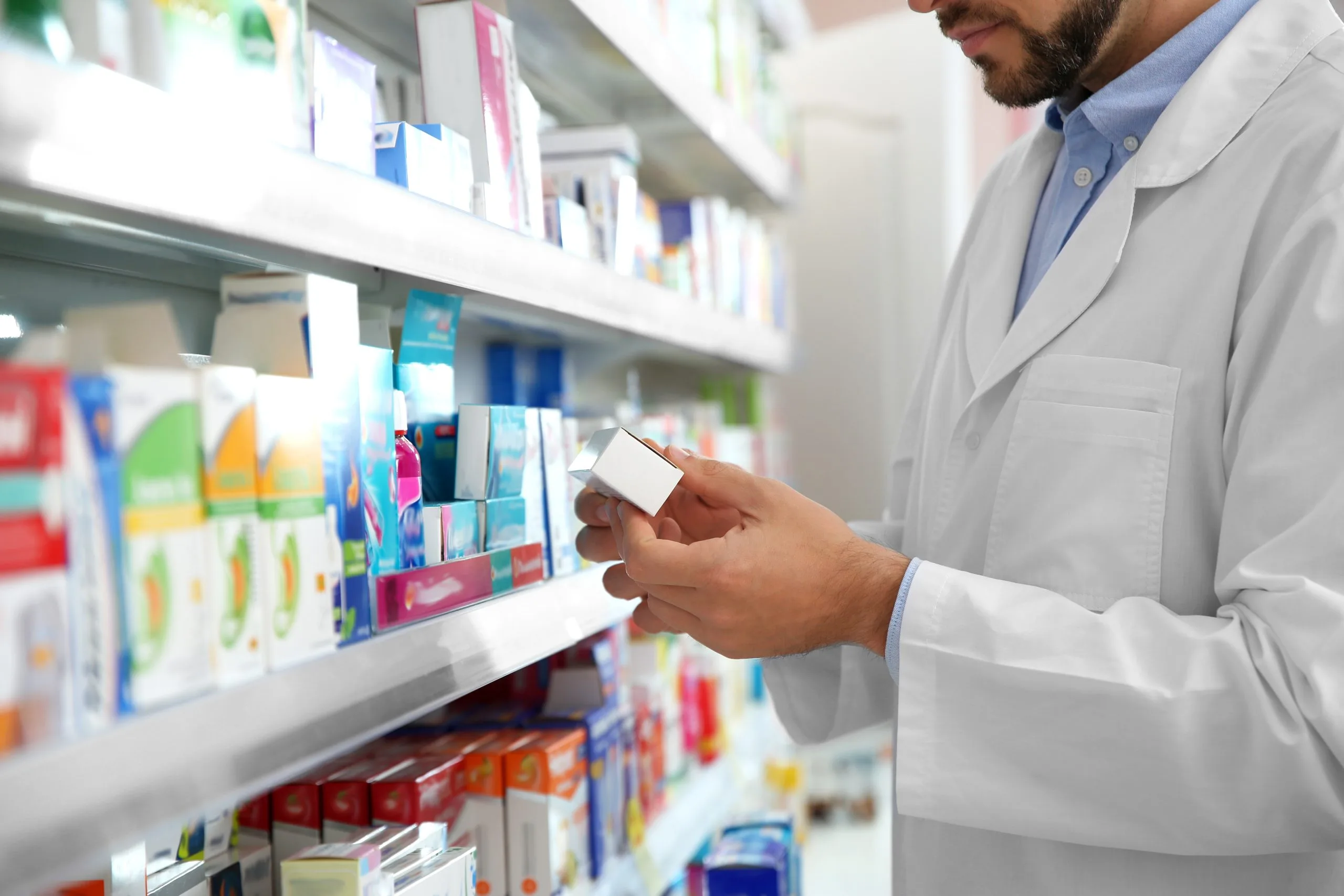 Pharmacist in white coat checking medication label while holding a bottle in front of pharmacy shelves