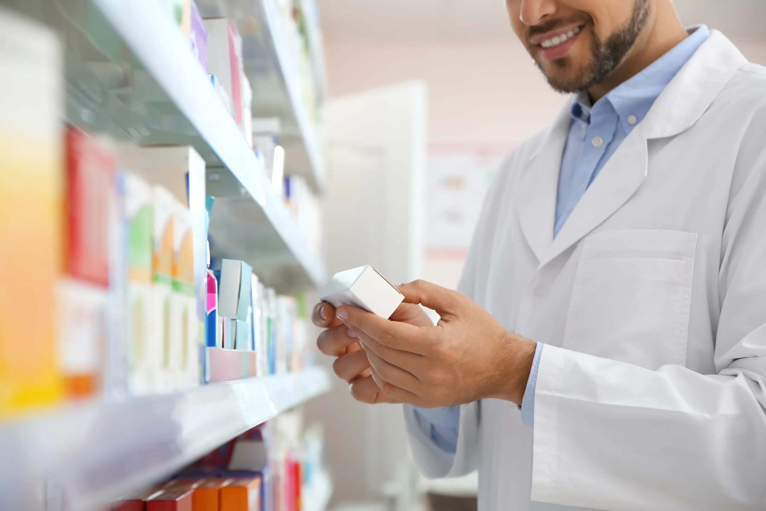 Pharmacist in white coat holding medication box while reviewing pharmacy shelves