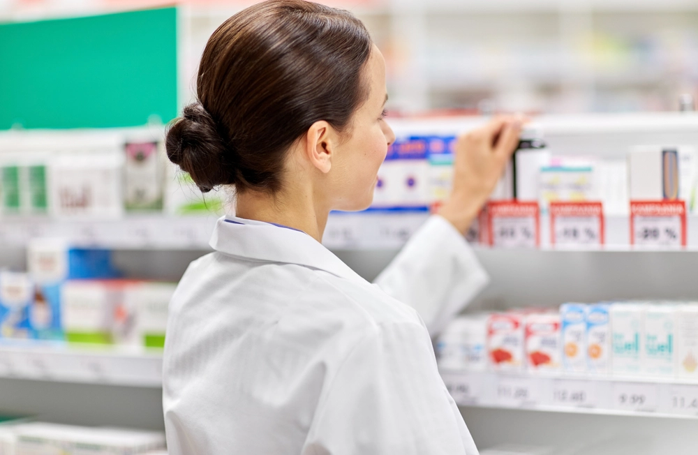 Female pharmacist in white coat reviewing medication inventory on pharmacy shelves