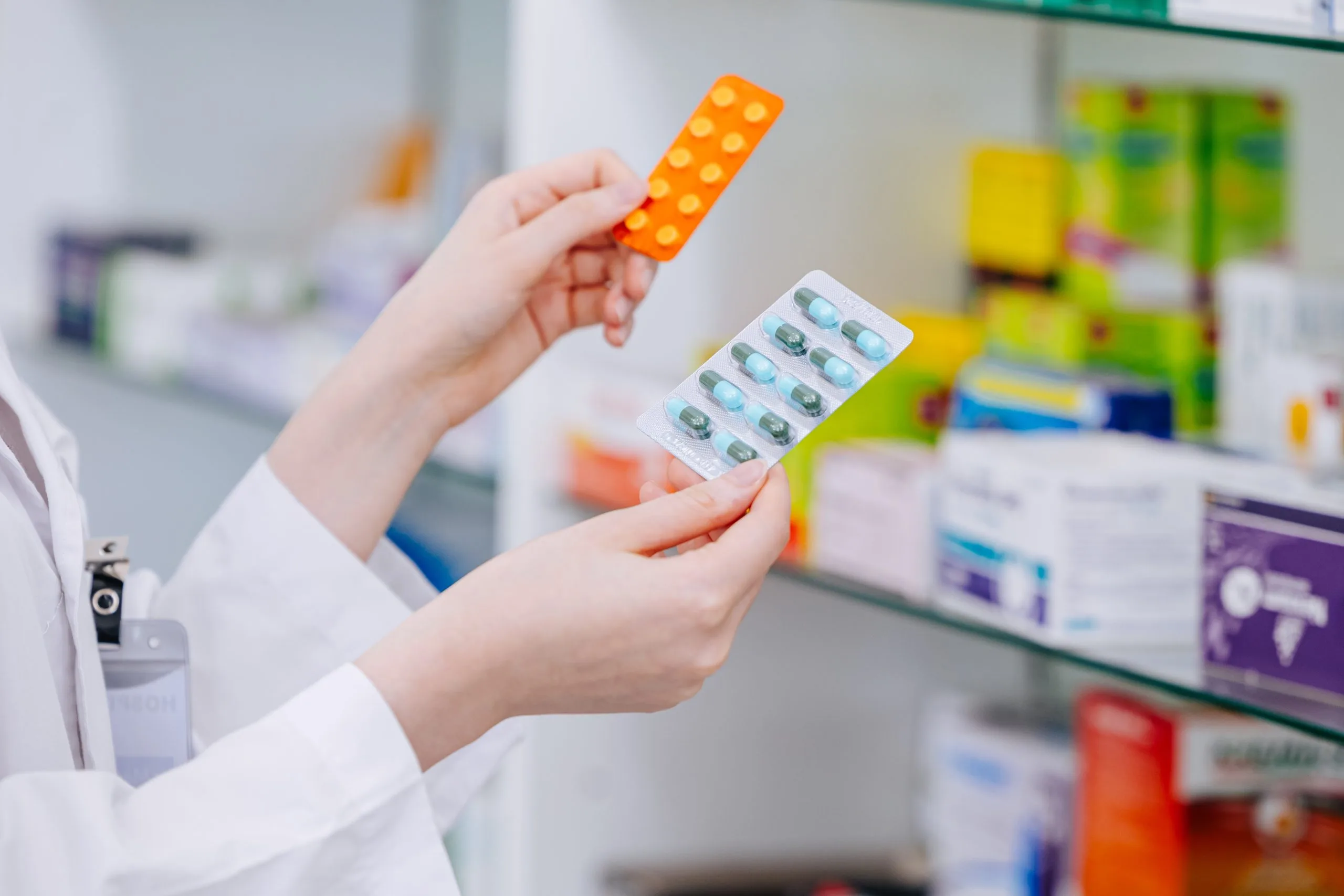 Pharmacist in white coat comparing orange and white medicine blister packs in front of pharmacy shelves