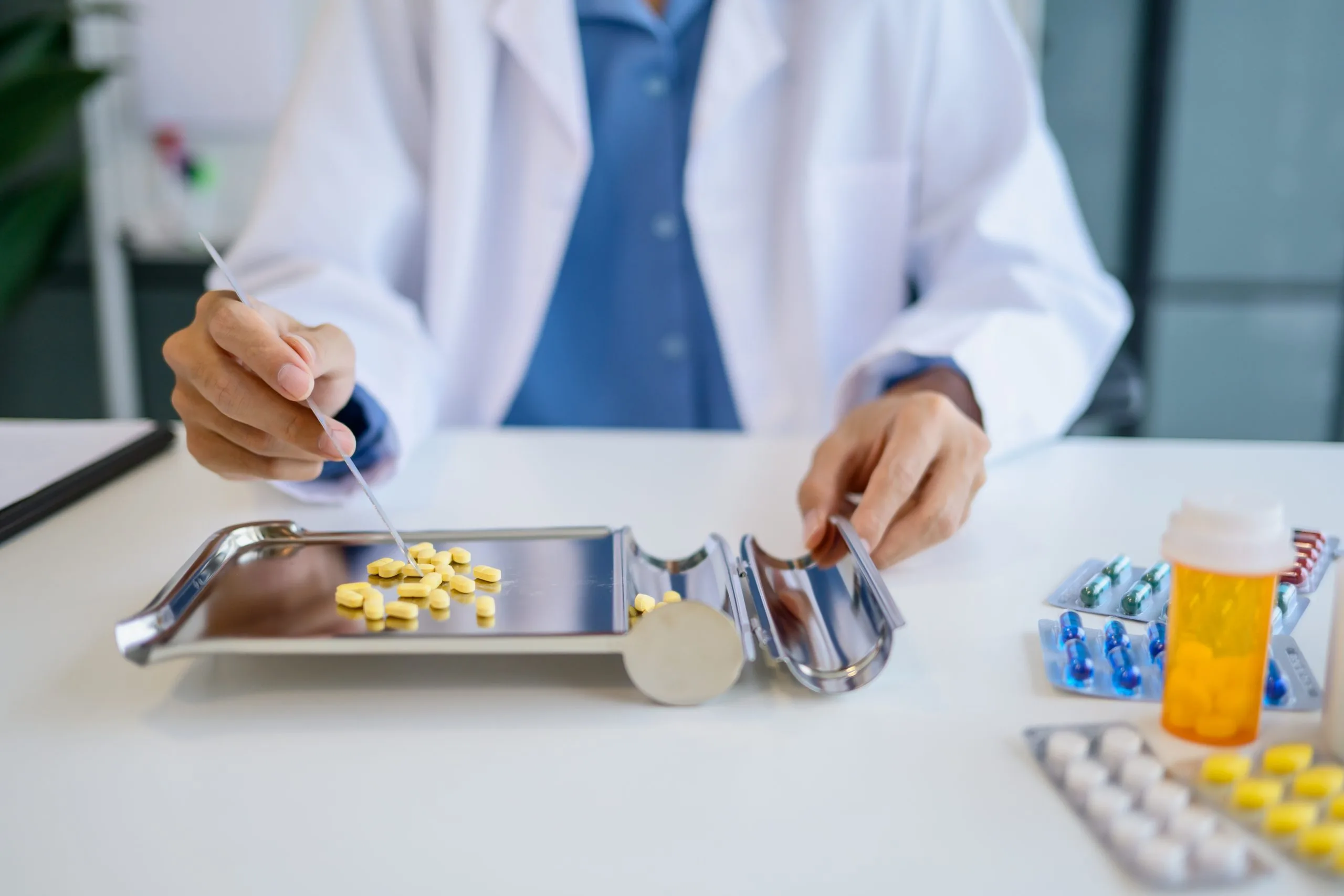 Pharmacist in white coat arranging yellow tablets on a metal tray with measuring tools and medication bottles