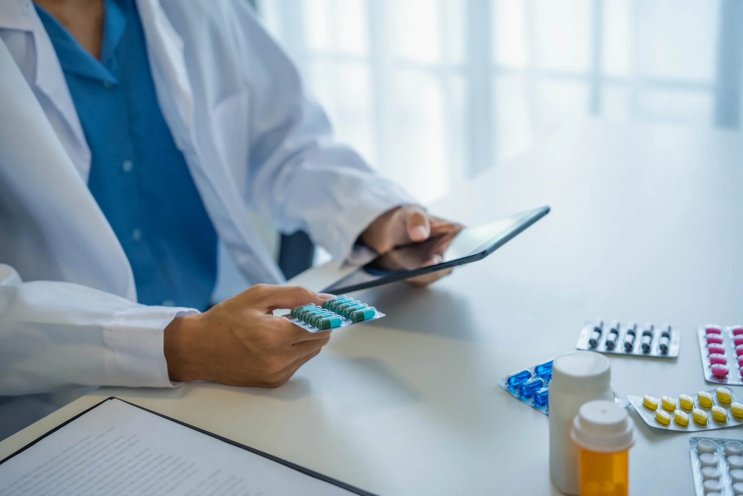 Pharmacist in white coat holding medication blister pack while using tablet to record dispensing information