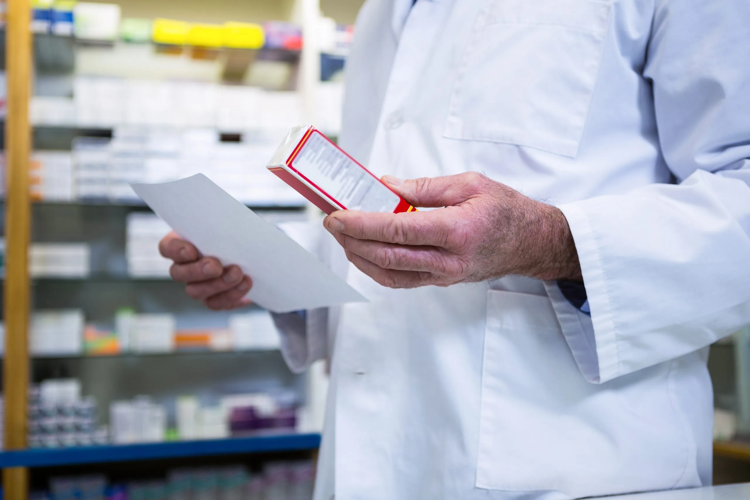 Pharmacist holding a prescription and medication box in a pharmacy with shelves of medicines in the background