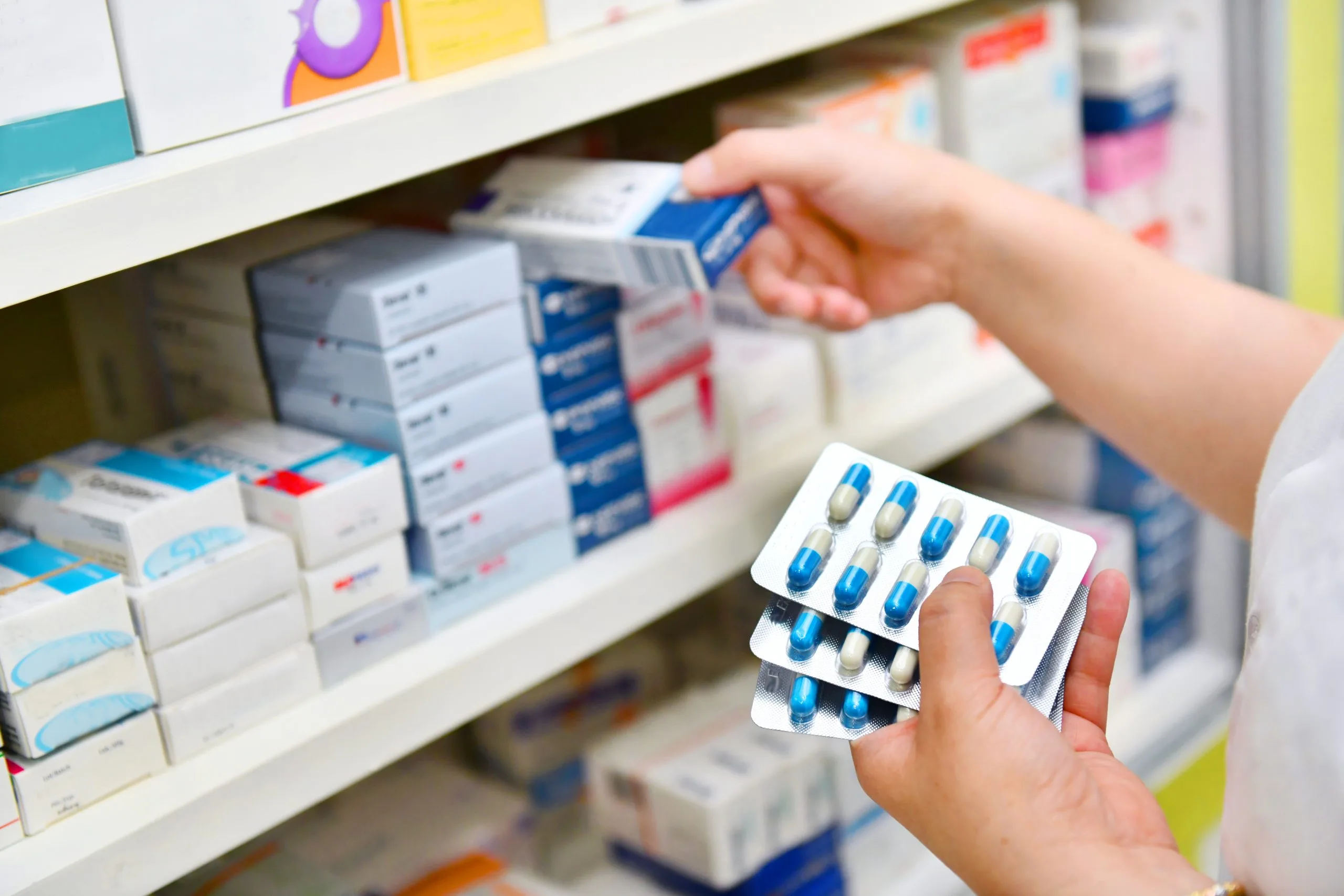 Pharmacist holding blue and white capsule blister packs while reaching for medication boxes on pharmacy shelf