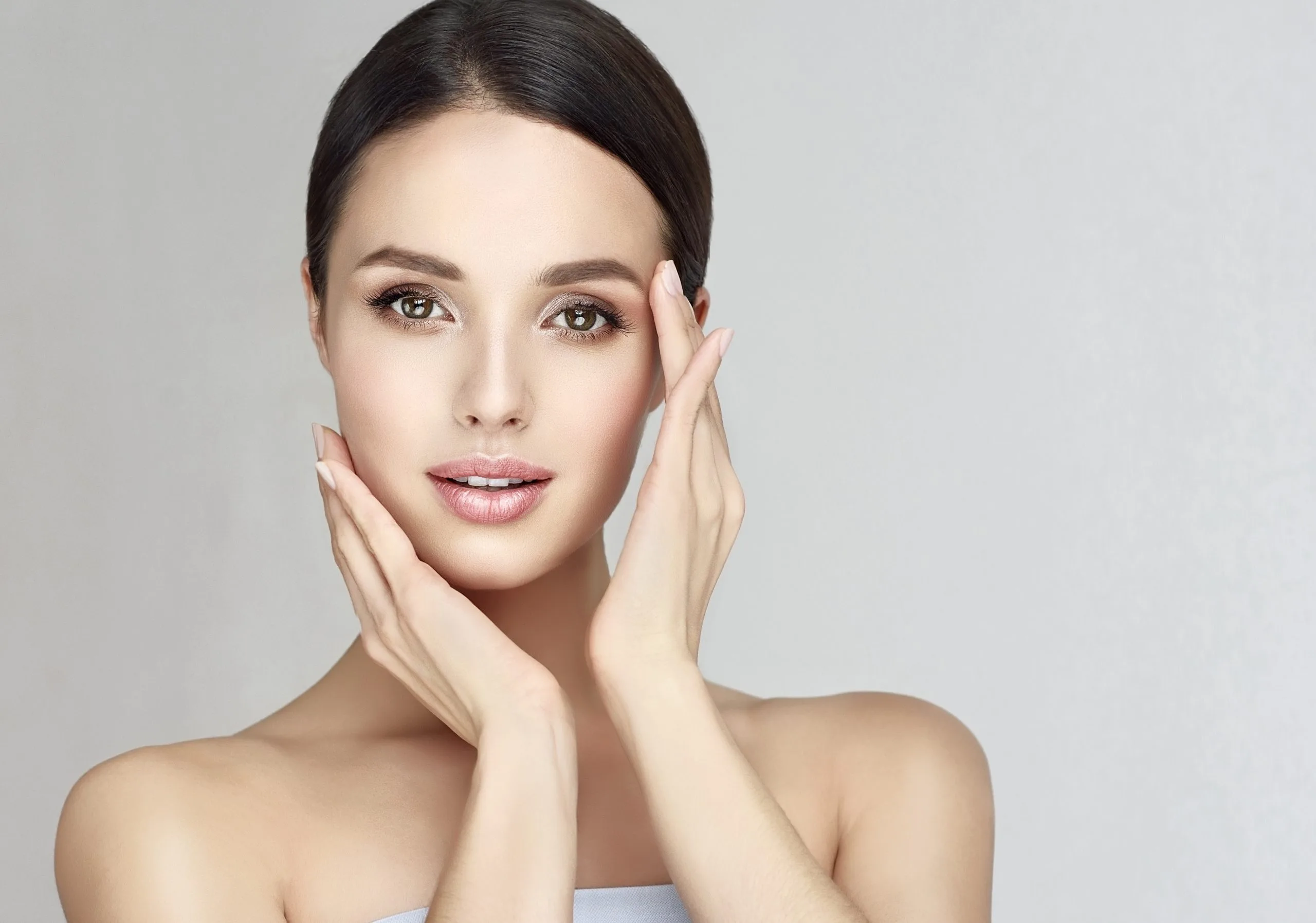 Woman with glowing skin touching her face with both hands against neutral background
