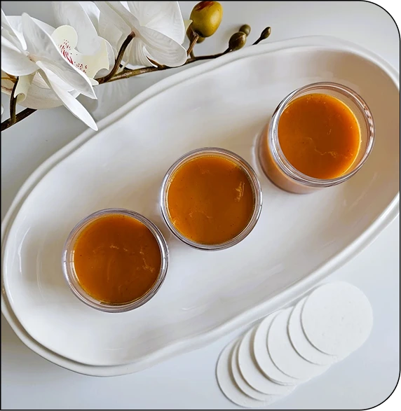 Three glass jars of amber glow bar serum on a white tray with white orchid and application pads
