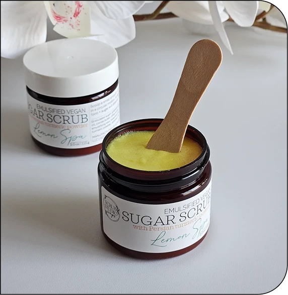 Two jars of CNT sugar scrub with wooden applicator, featuring lemon and vegan formulas