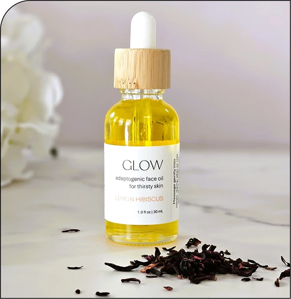 GLOW adaptogenic face oil bottle with dropper cap and hibiscus petals on white surface