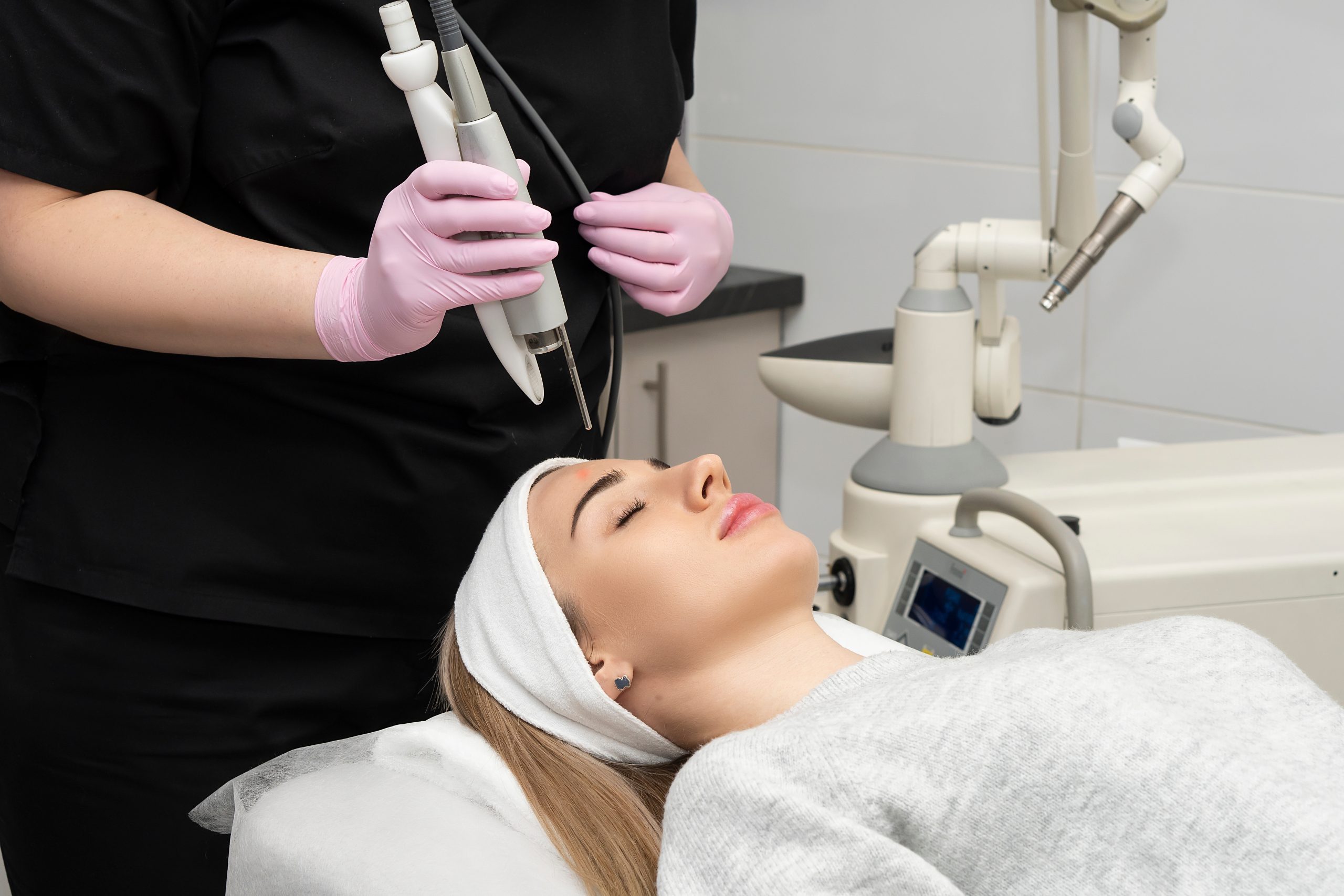 Practitioner in pink gloves holding laser handpiece above patient's face during treatment in medical spa setting
