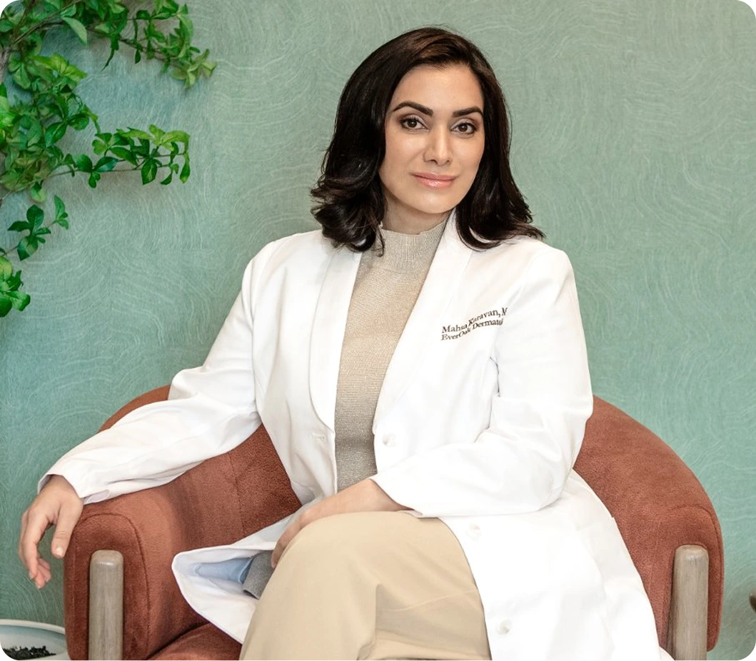 Mahsa Karavan, MD in white coat, seated in modern clinic setting with green wall and potted plant