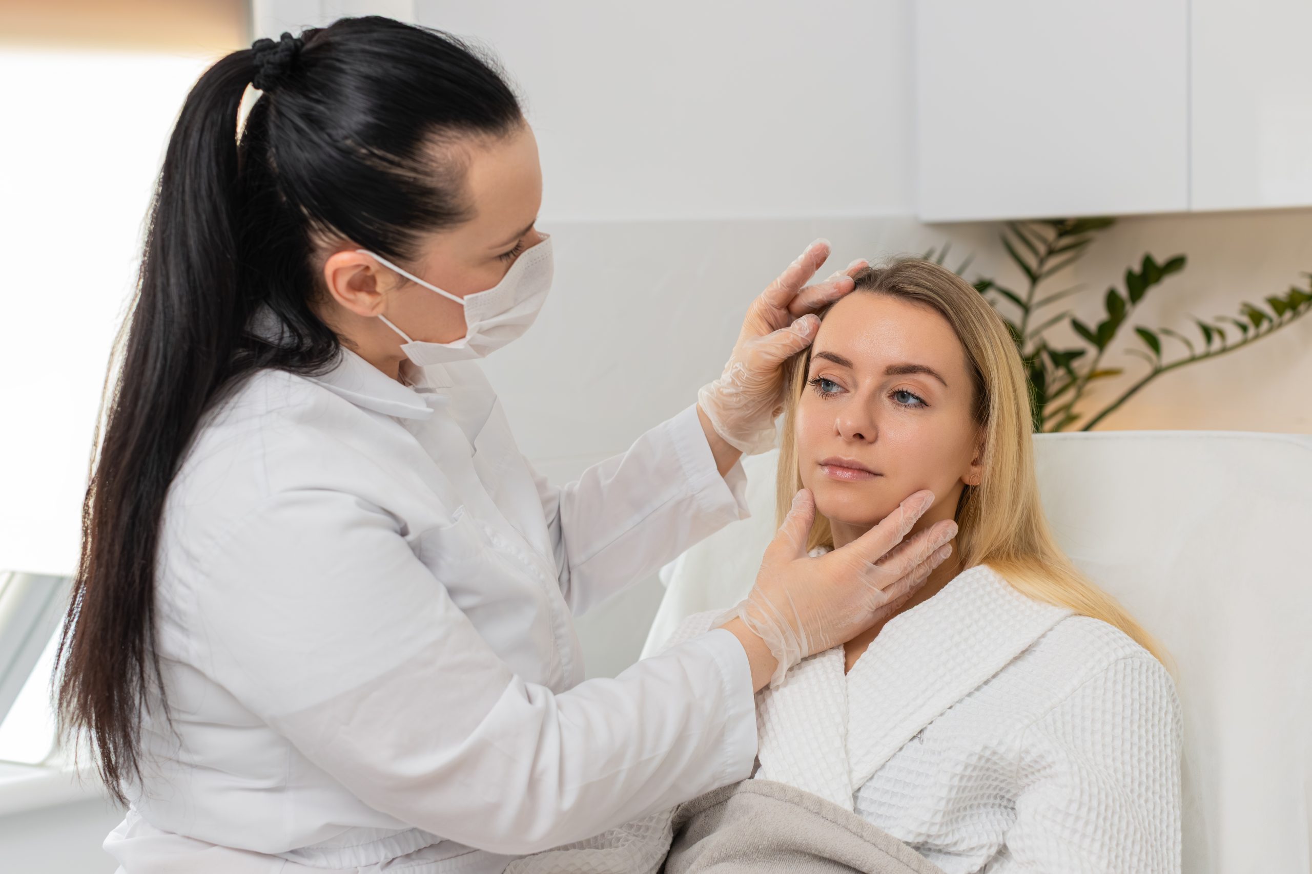 Clinician in mask examining patient's face and jawline during consultation