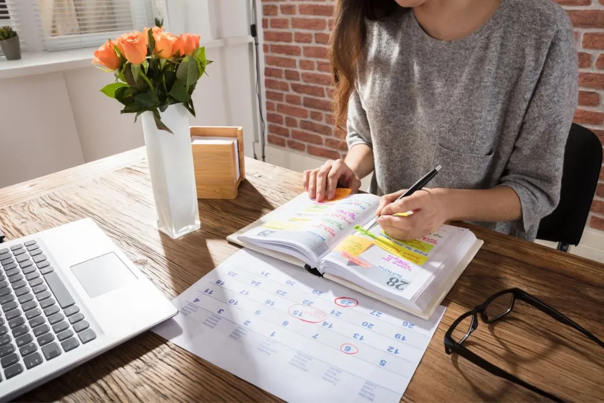 Woman at a wooden desk color-coding tasks in a paper planner beside her laptop and a monthly calendar