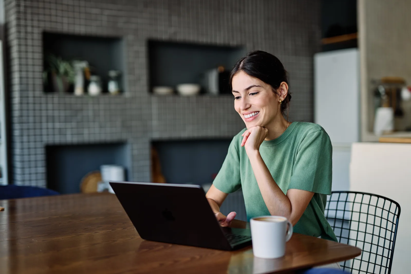 Smiling woman sitting at a kitchen table with a coffee mug, working on her laptop at home