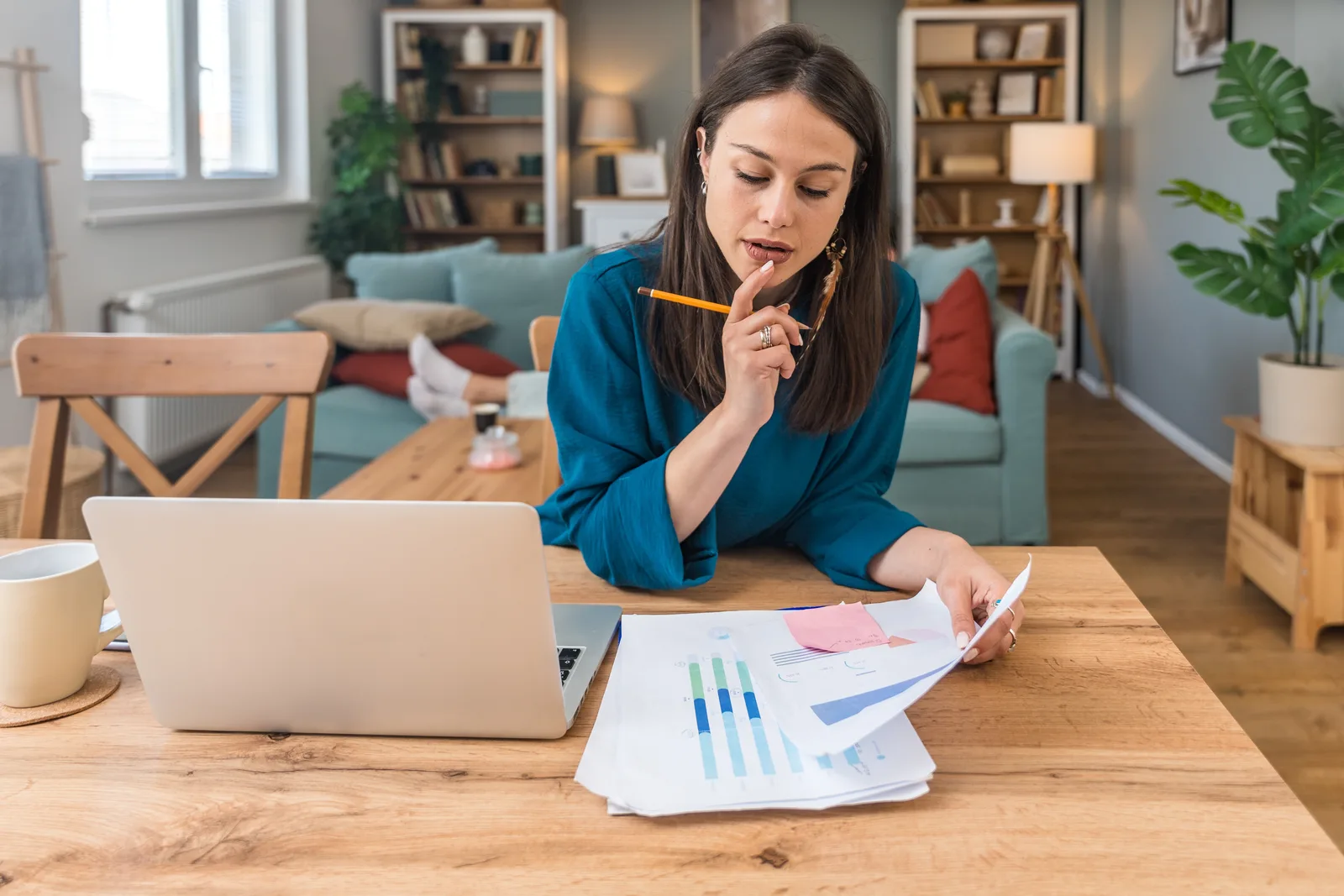 Woman sitting at a wooden table at home thinking through charts and papers next to her laptop