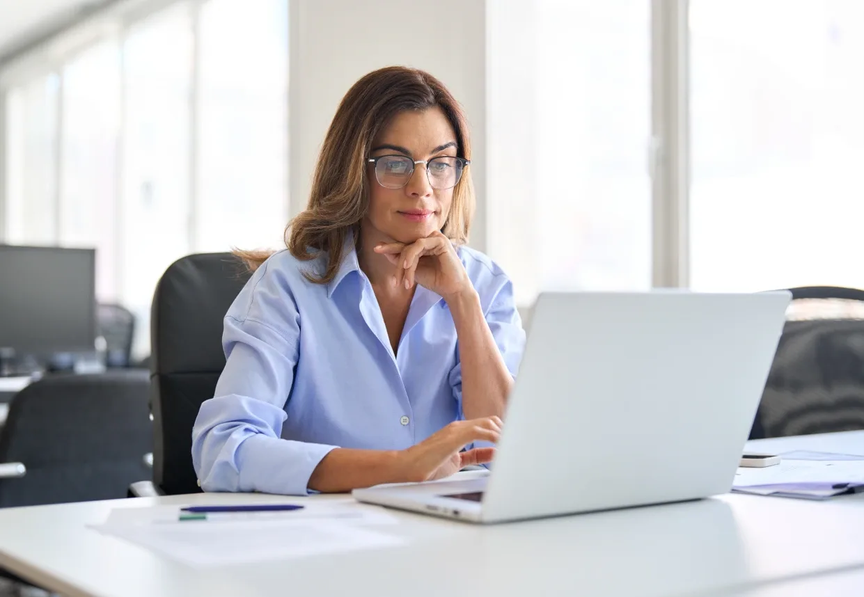 Professional woman in glasses sitting at a desk in a bright office, thinking while working on her laptop