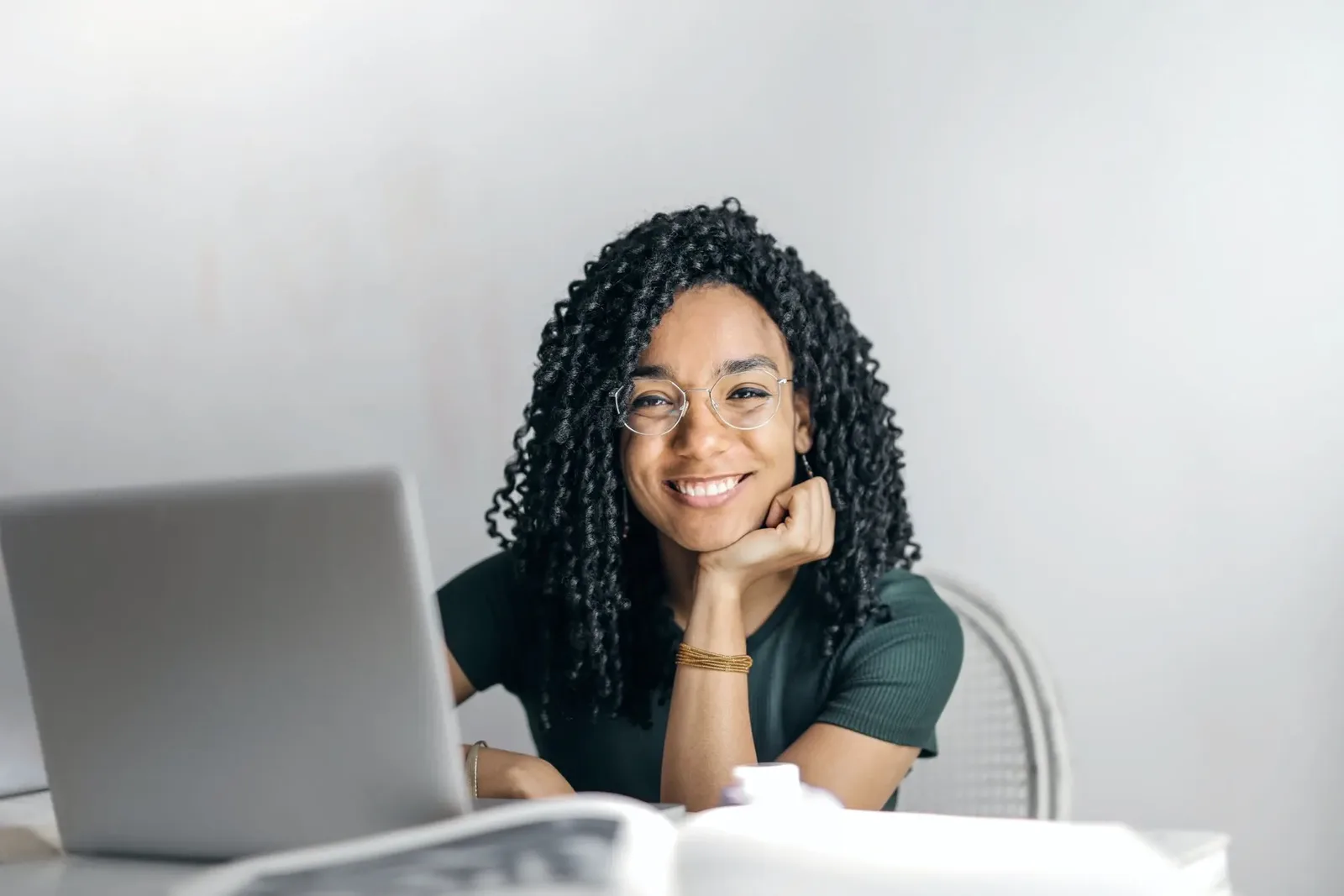 Smiling young woman with curly hair and glasses sitting at a desk with her laptop and notebook