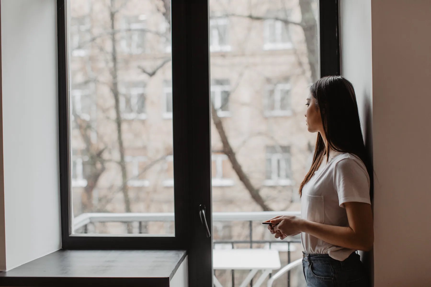 Woman by a window holding her phone, looking away in quiet reflection
