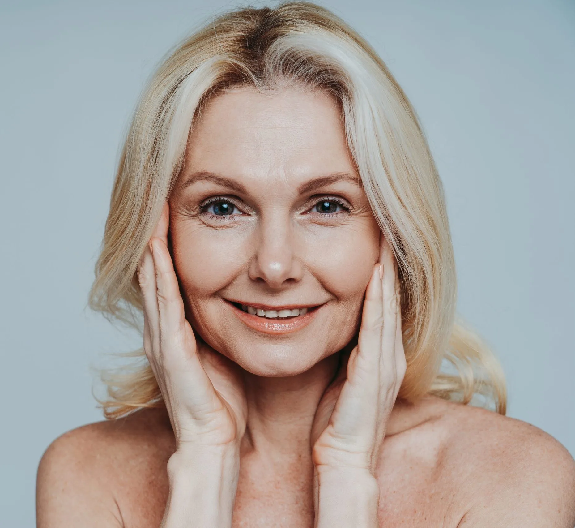 Mature woman with blonde hair smiling and touching her face with both hands against a blue background