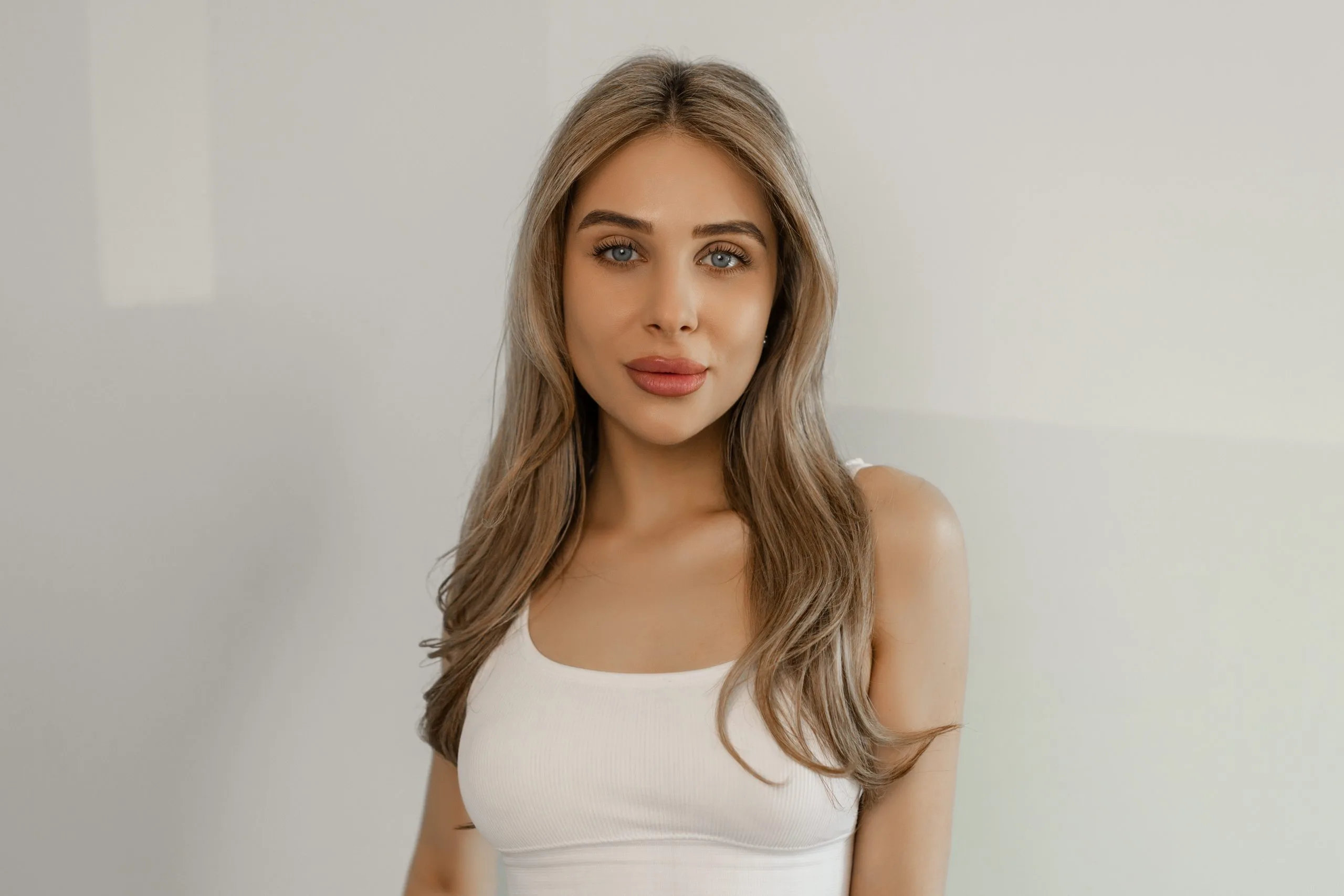 Woman with long brown hair and full lips wearing a white sleeveless top against a neutral background