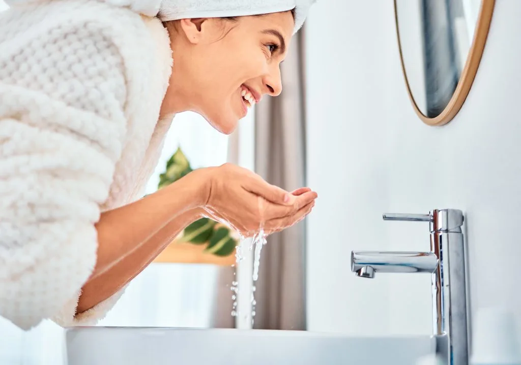 Woman in towel and headwrap washing her face with water at bathroom sink