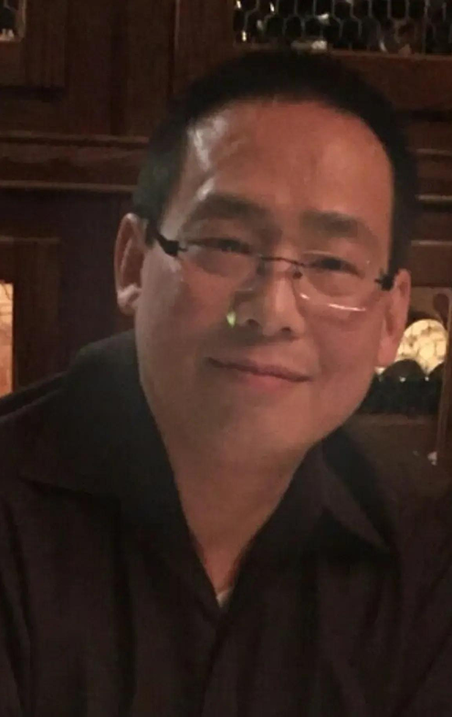 Man with short dark hair and glasses wearing a dark shirt, smiling indoors