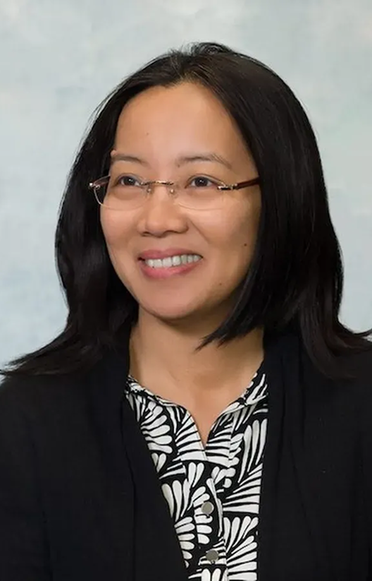 Woman with long black hair and glasses wearing a black blazer and patterned blouse, smiling at camera