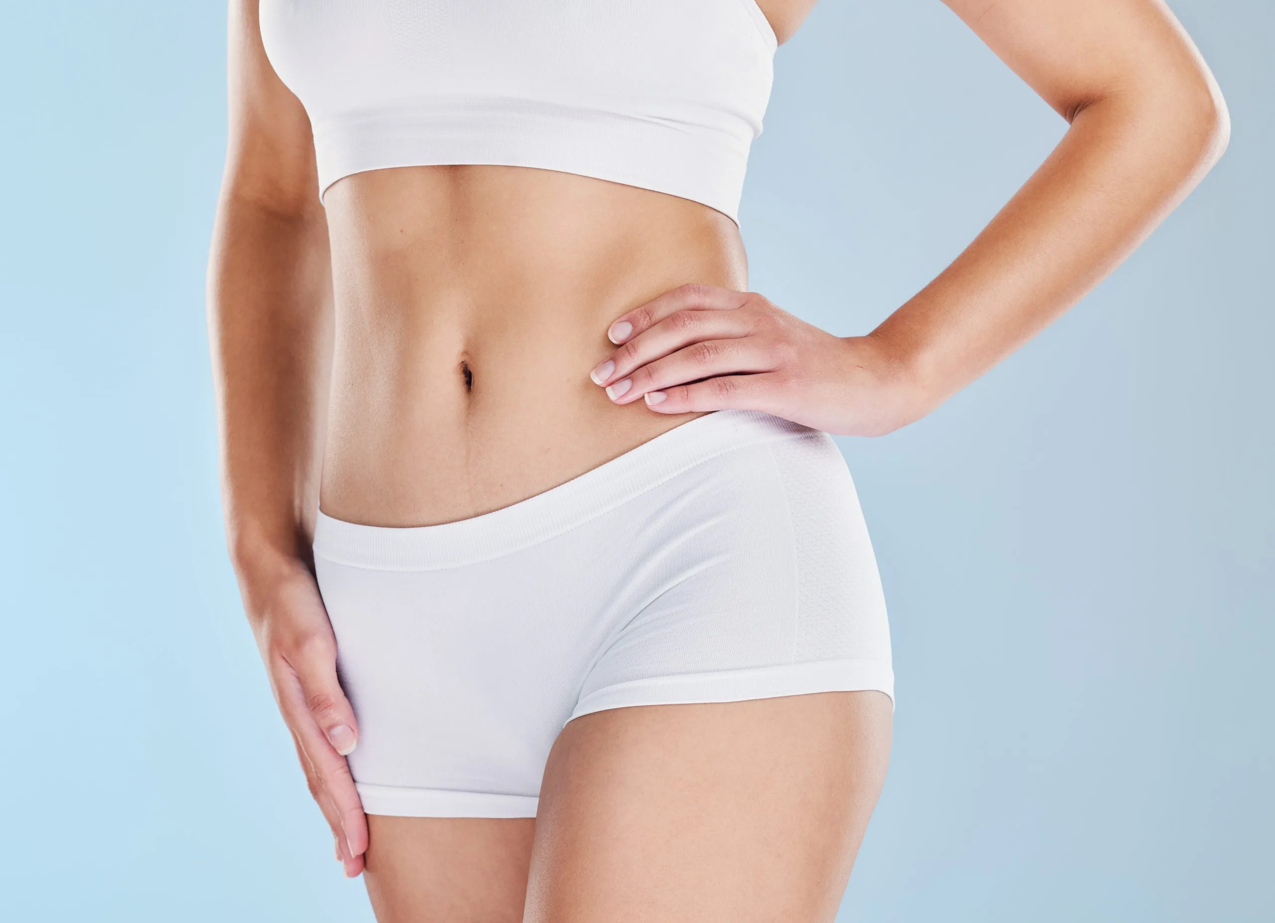 Woman in white crop top and shorts touching her toned abdomen against a blue background