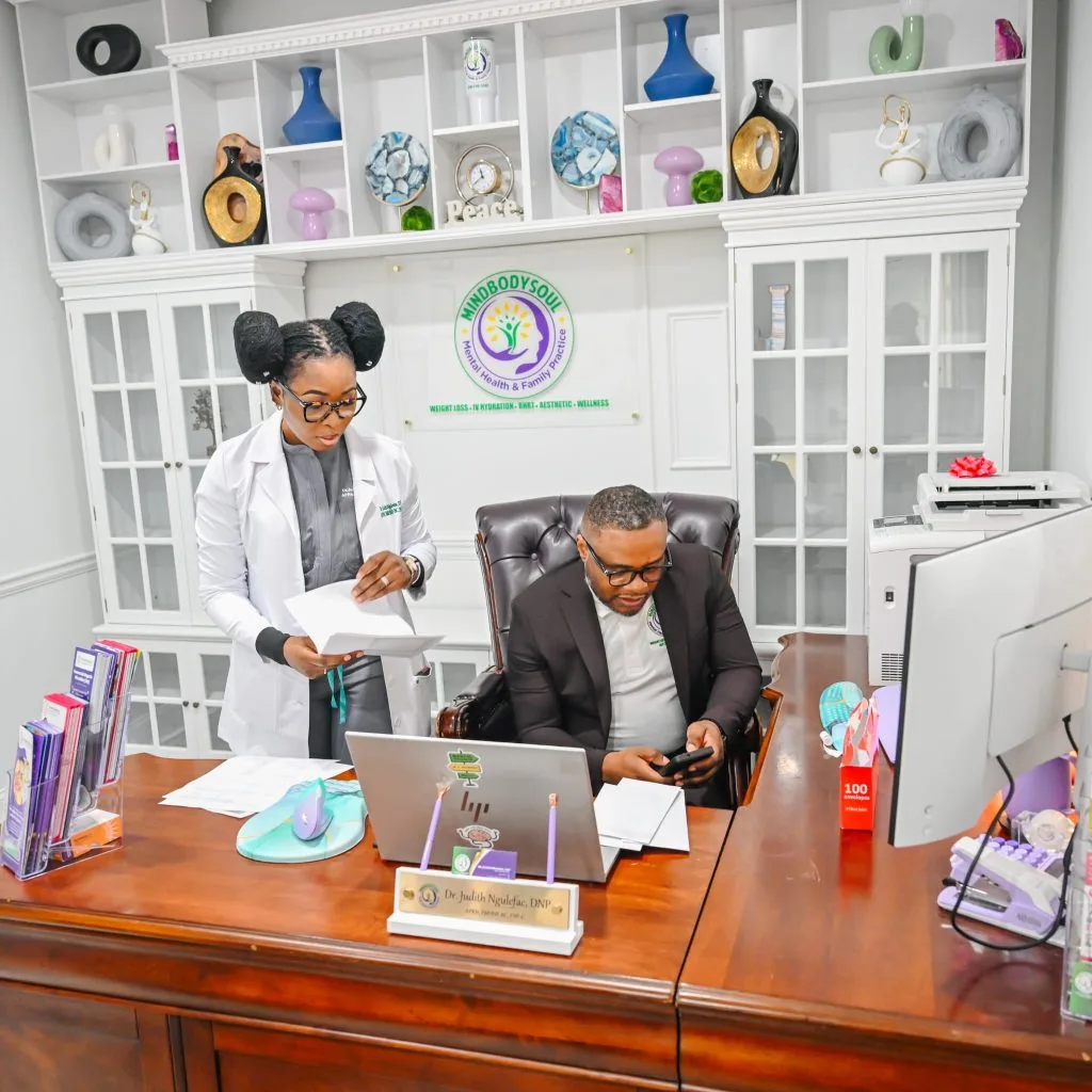 Dr. Judith Ngulefac and a colleague review documents at a desk in MindBodySoul's modern office with the practice logo displayed on the wall behind them
