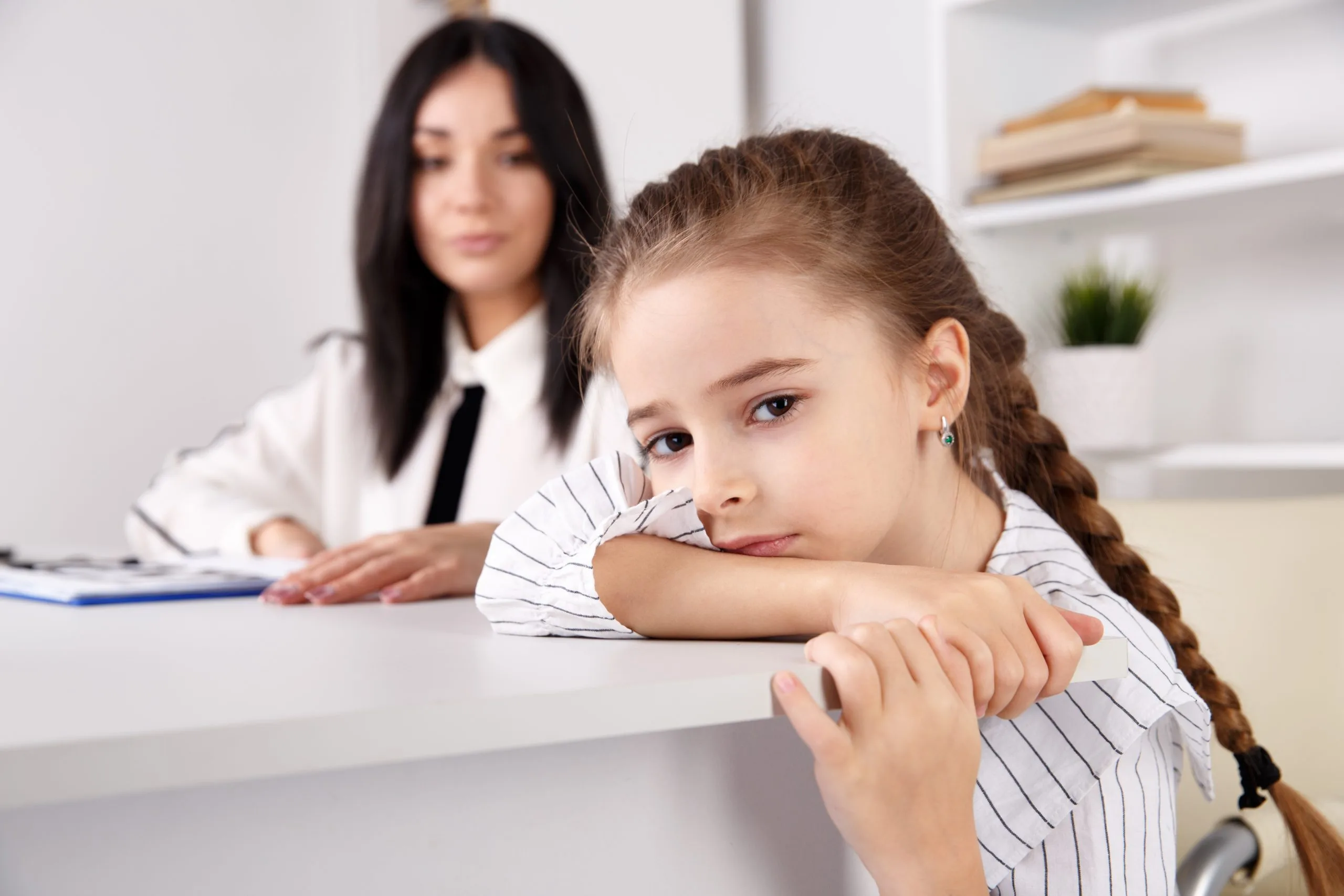 Young girl leaning on desk looking sad or disengaged with woman in white coat behind her