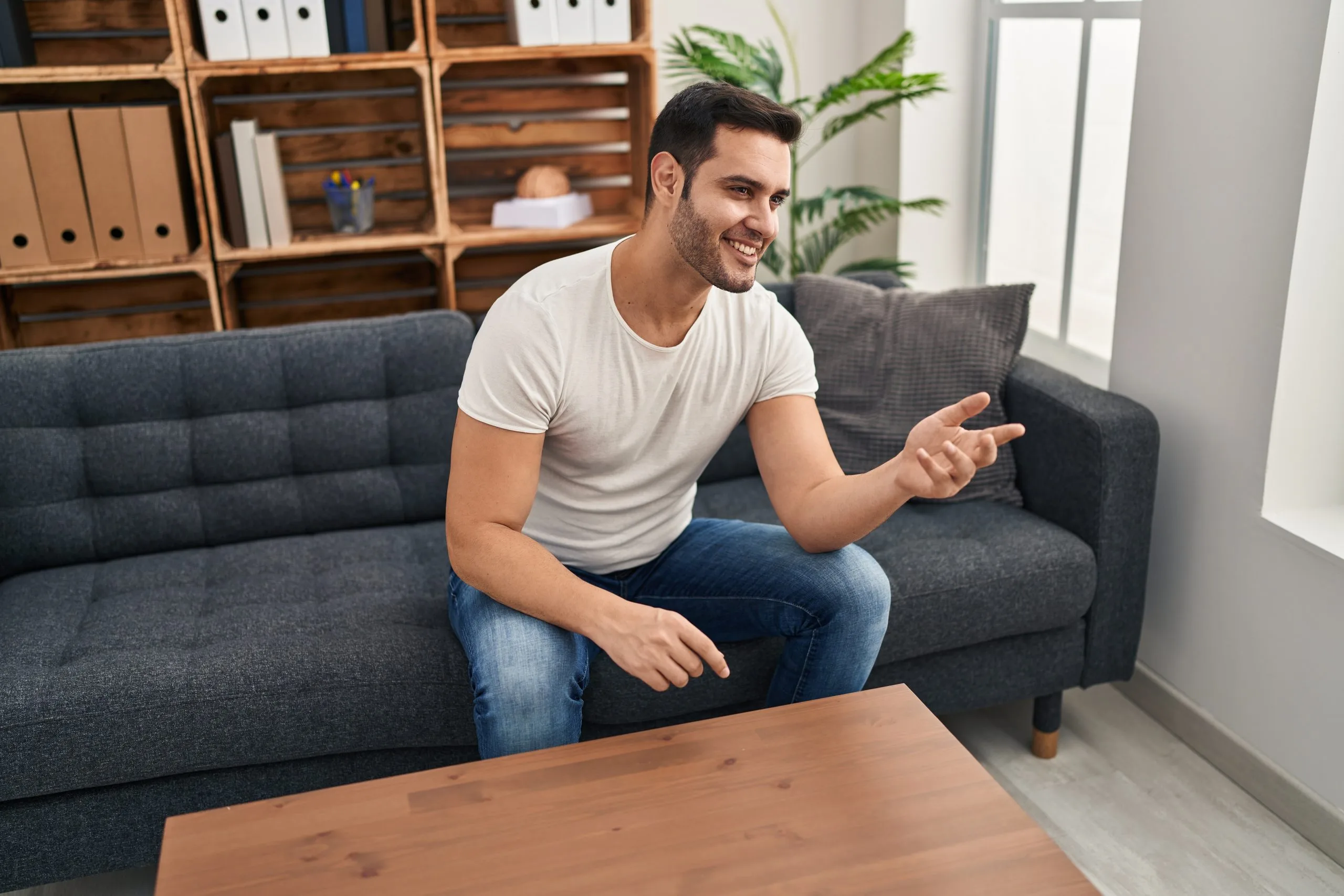 Man sitting on gray sofa in living room with wooden shelving, gesturing while speaking