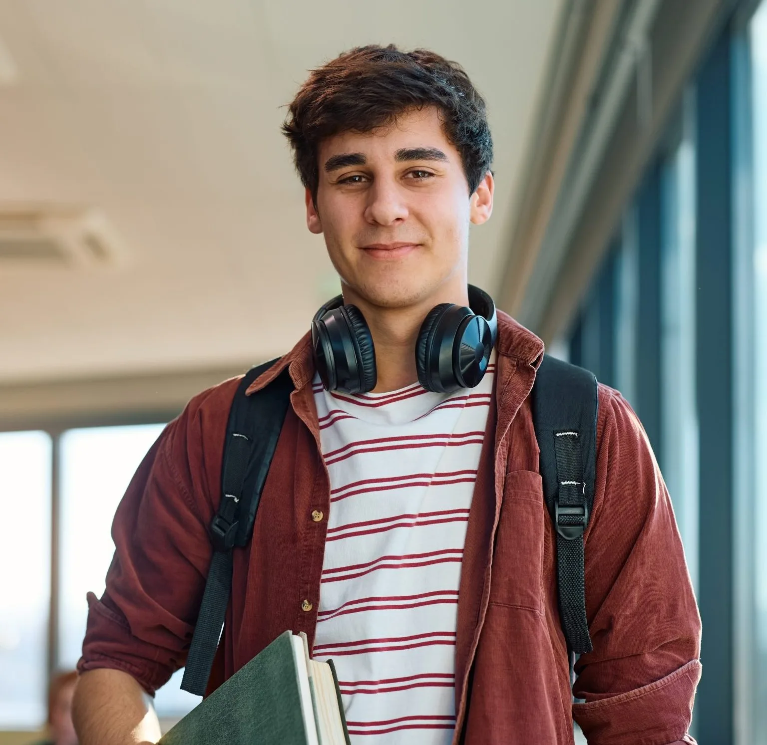 Smiling teenager wearing headphones around neck, backpack, and striped shirt holding book